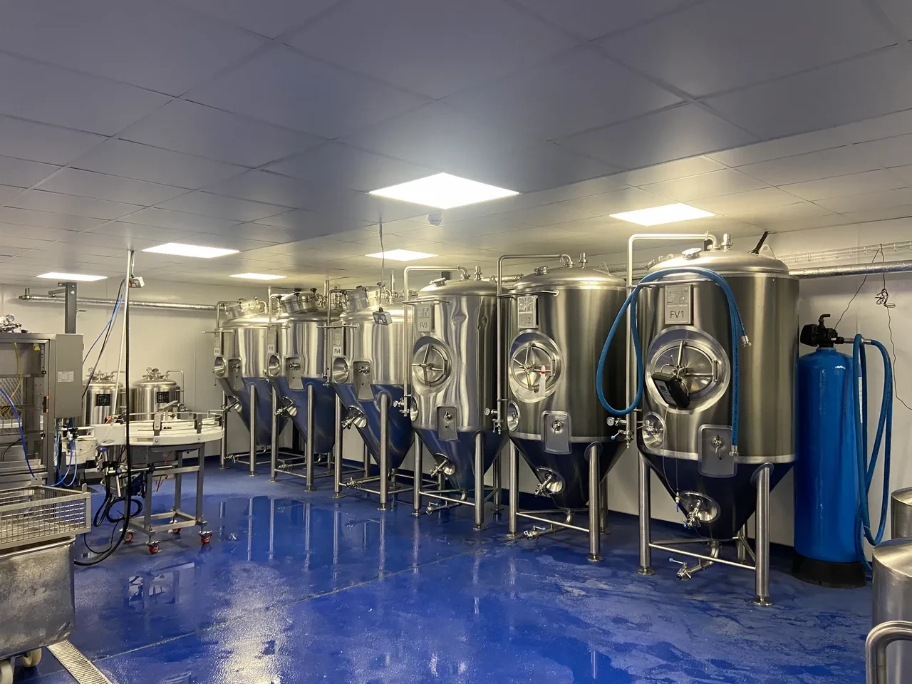 Row of large stainless steel fermentation tanks in a clean industrial brewery room with blue floor.