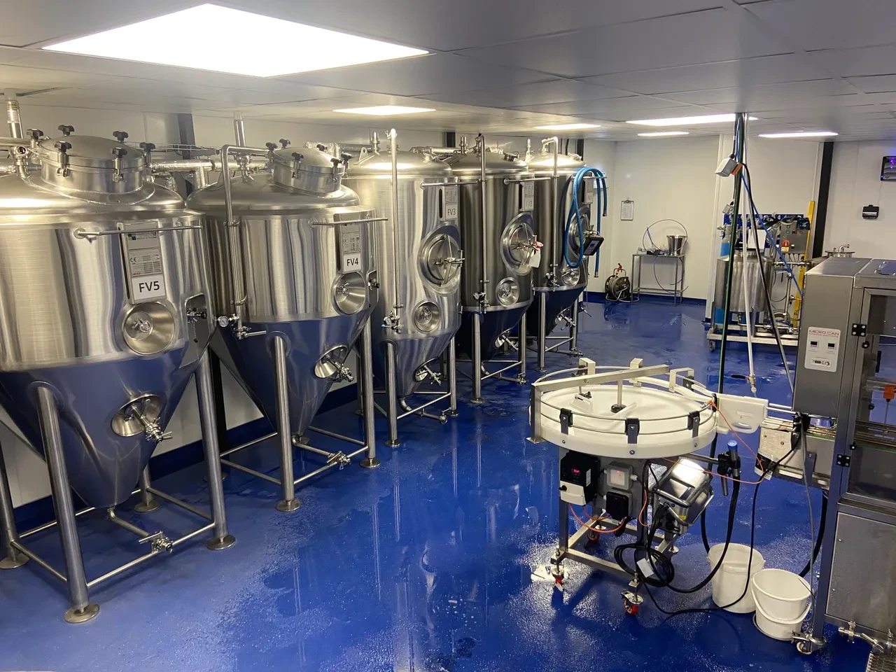 A clean brewery room with five large labeled stainless steel fermentation tanks and packaging equipment on blue floor.