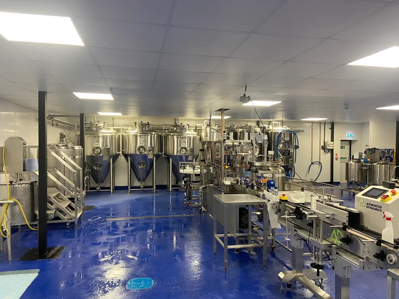 Spacious industrial brewery with stainless steel tanks, machines, and wet blue floors under bright lights.